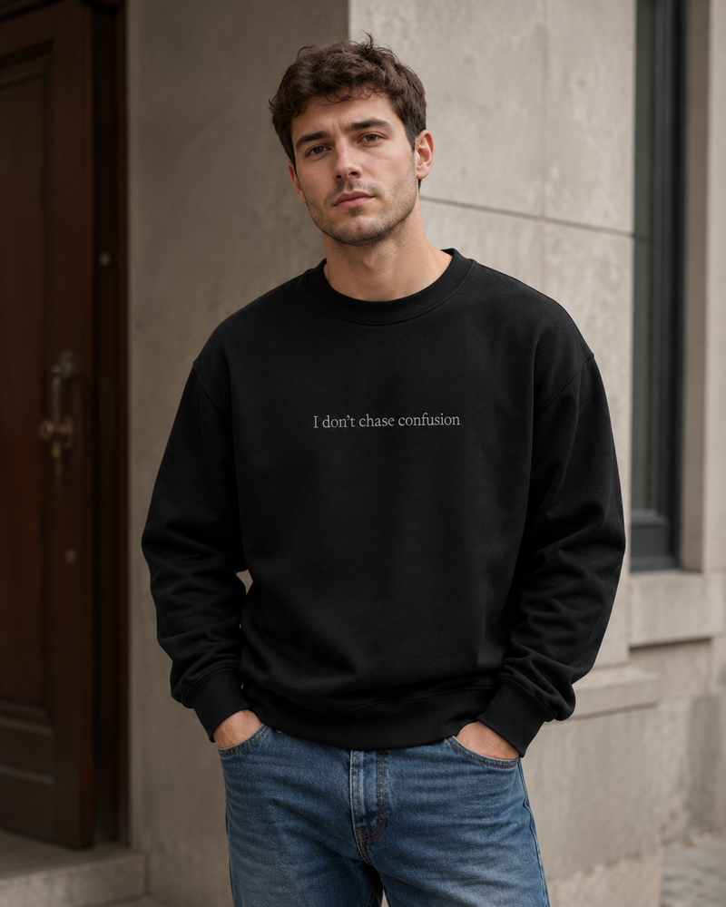 A man wearing a black crewneck sweatshirt with the text 'I don't chase confusion' printed in white across the chest, paired with blue jeans, standing outdoors against a stone wall.