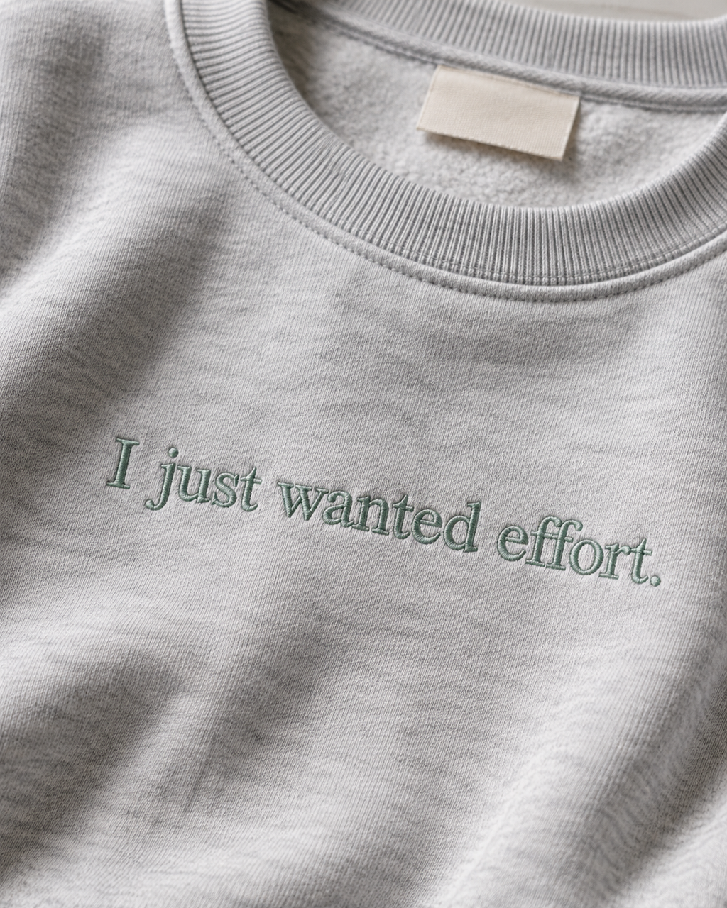 Folded Heather Gray crewneck sweatshirt with dark embroidery reading “I just wanted effort.” on a light background.