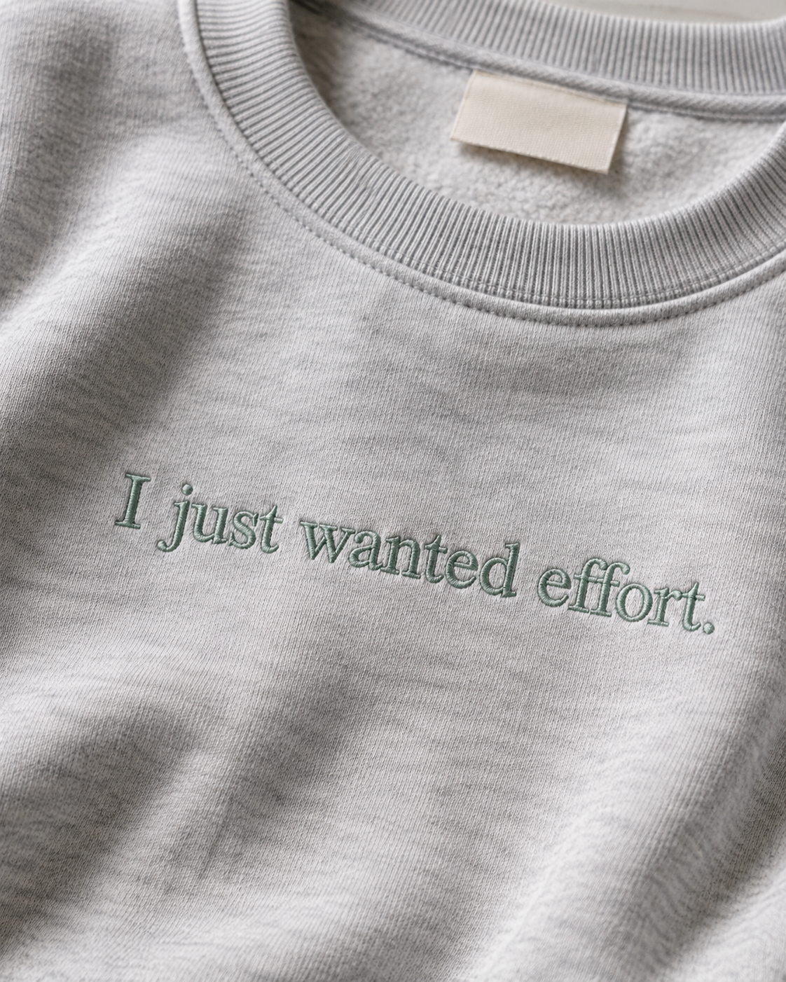 Folded Heather Gray crewneck sweatshirt with dark embroidery reading “I just wanted effort.” on a light background.