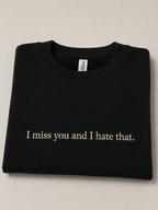 A folded black crewneck sweatshirt with the text 'I miss you and I hate that.' printed in white serif font across the chest, displayed on a neutral light beige background.
