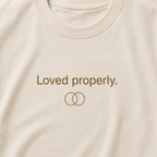 Close-up of a cream-colored t-shirt featuring the brown text 'Loved properly.' and a simple interlocking rings graphic on the chest.