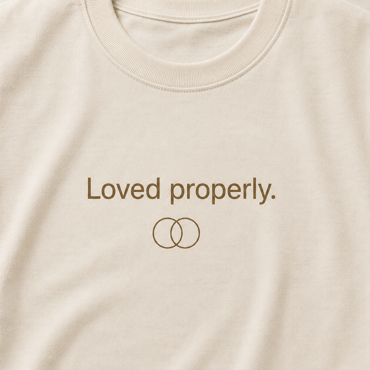 Close-up of a cream-colored t-shirt featuring the brown text 'Loved properly.' and a simple interlocking rings graphic on the chest.