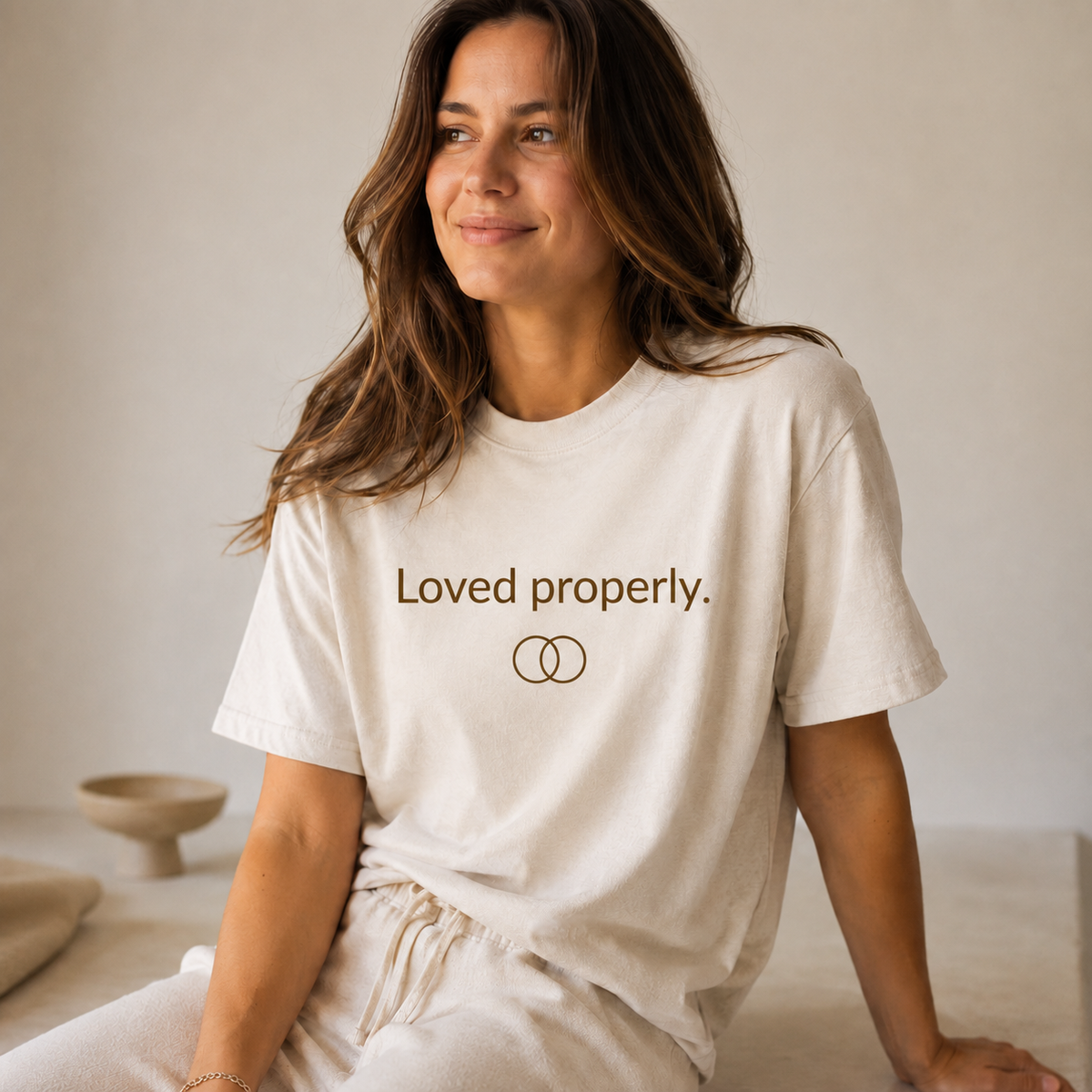 Warm cream T-shirt with camel brown text reading “Loved properly.” above two interlocking circles.