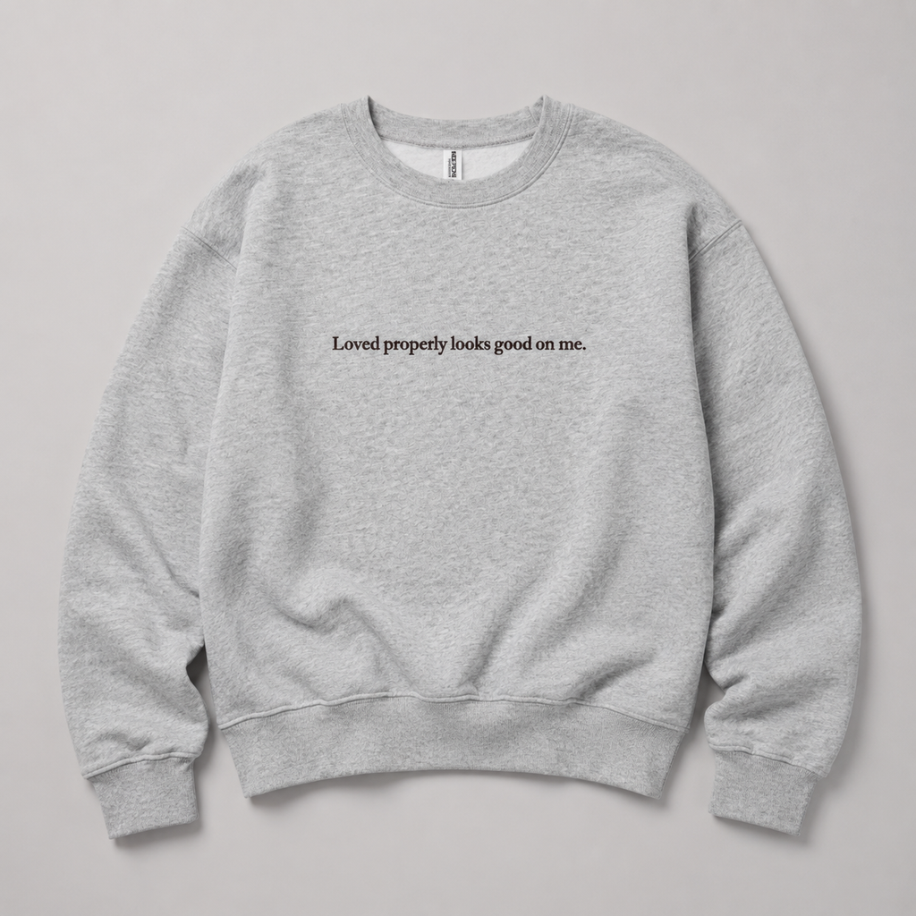 A light heather gray crewneck sweatshirt with the black printed text 'Loved properly looks good on me.' across the chest, displayed flat against a neutral gray background.