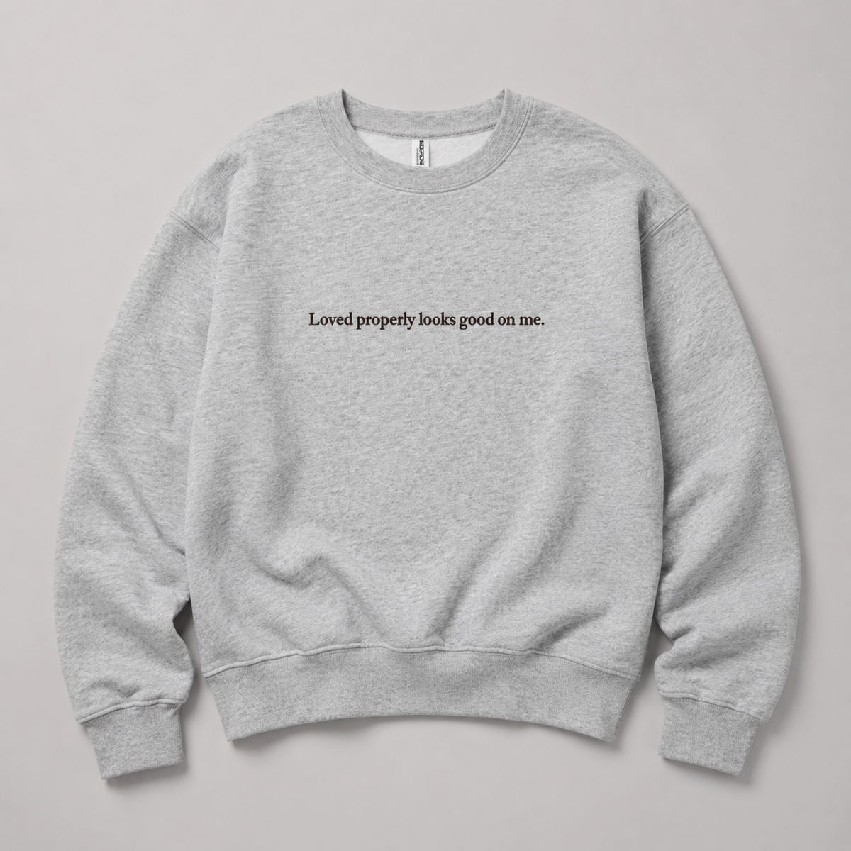 A light heather gray crewneck sweatshirt with the black printed text 'Loved properly looks good on me.' across the chest, displayed flat against a neutral gray background.