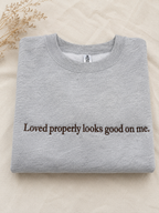 A folded light gray crewneck sweatshirt with the phrase 'Loved properly looks good on me.' printed in dark brown serif text across the chest, displayed on a beige fabric background with dried floral stems in the corner.