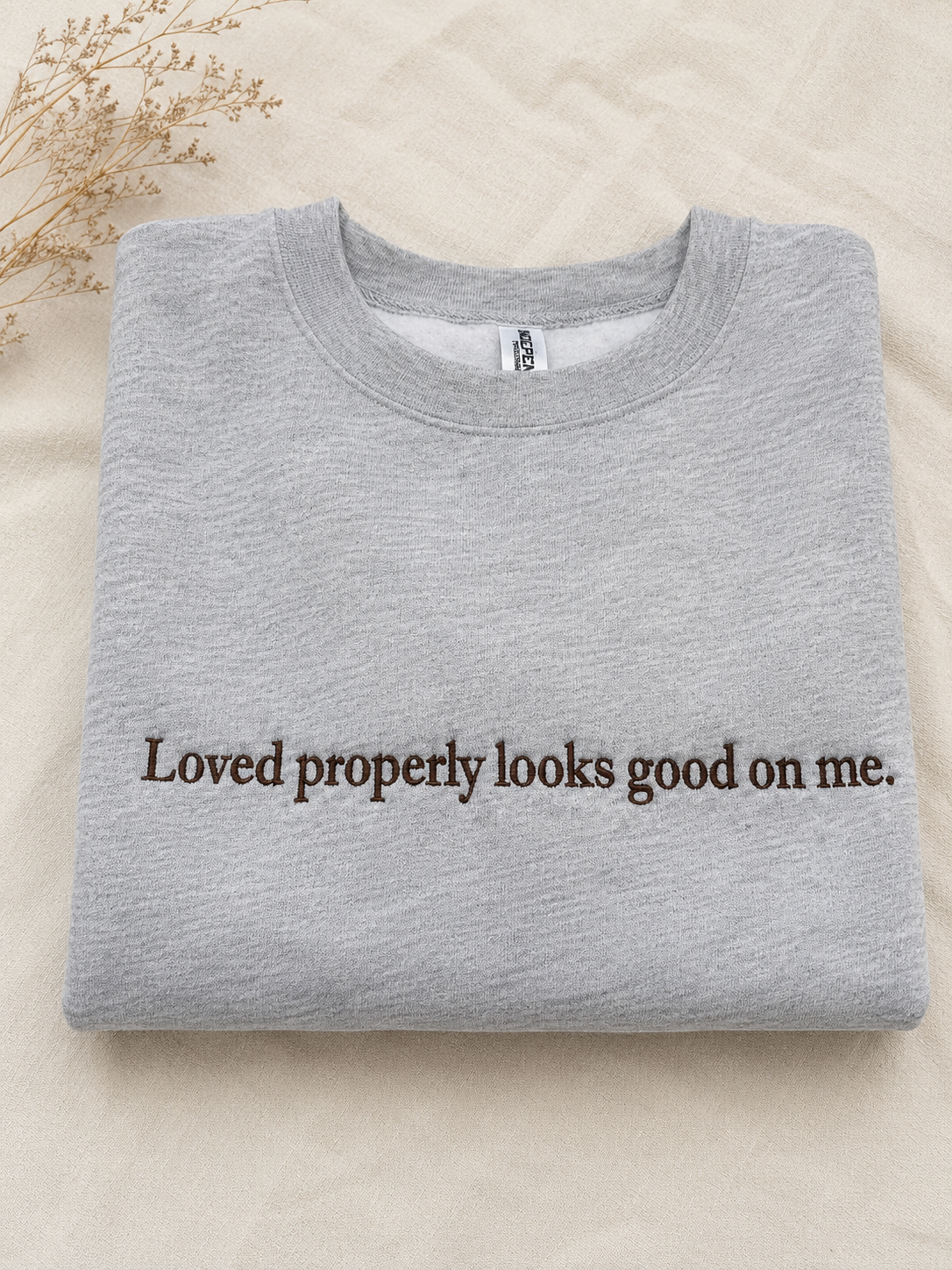 A folded light gray crewneck sweatshirt with the phrase 'Loved properly looks good on me.' printed in dark brown serif text across the chest, displayed on a beige fabric background with dried floral stems in the corner.