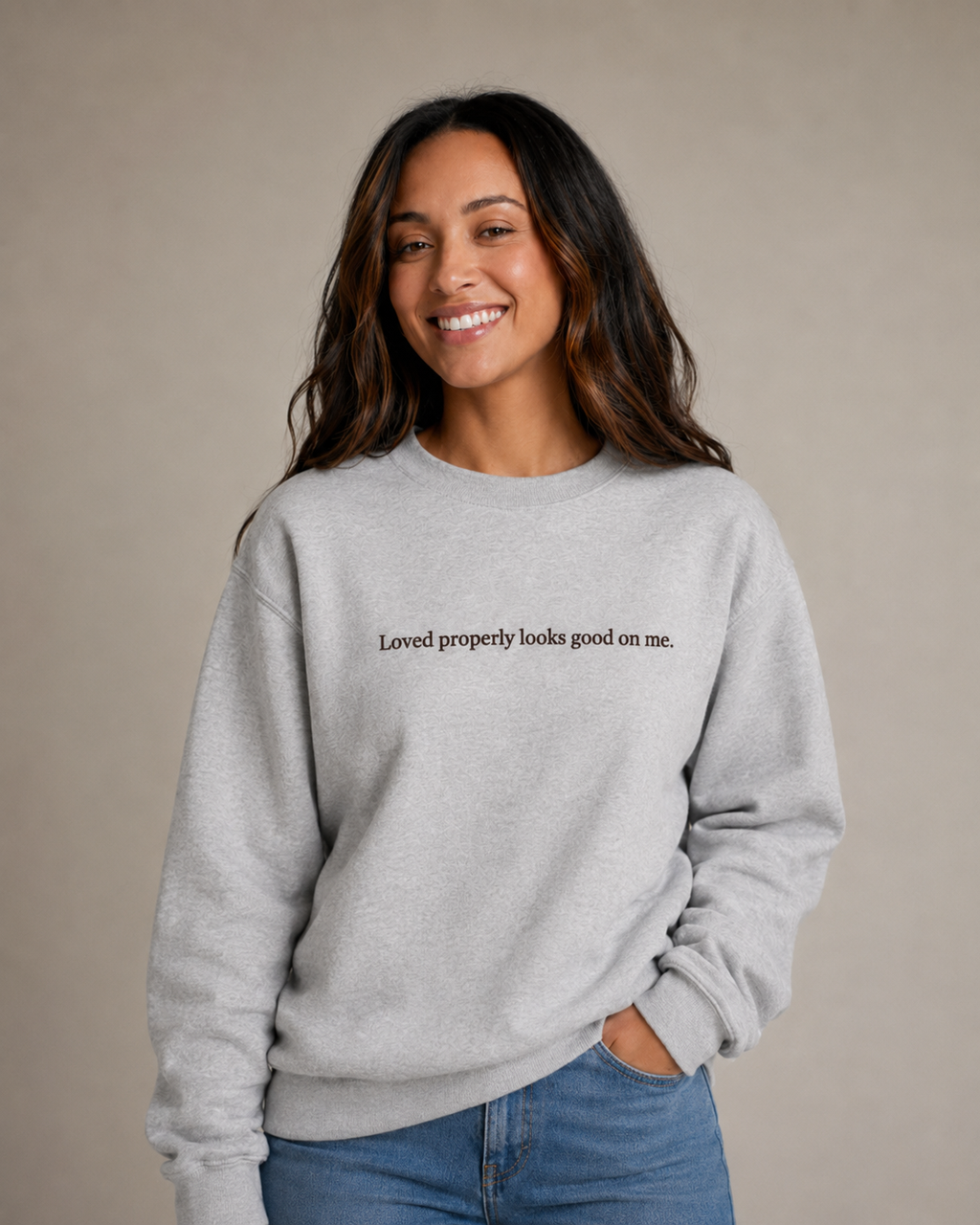 Folded Heather Gray crewneck sweatshirt with dark chocolate embroidery reading “Loved properly looks good on me.” on a light beige fabric background.