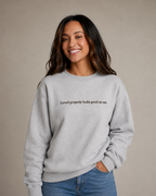 Folded Heather Gray crewneck sweatshirt with dark chocolate embroidery reading “Loved properly looks good on me.” on a light beige fabric background.