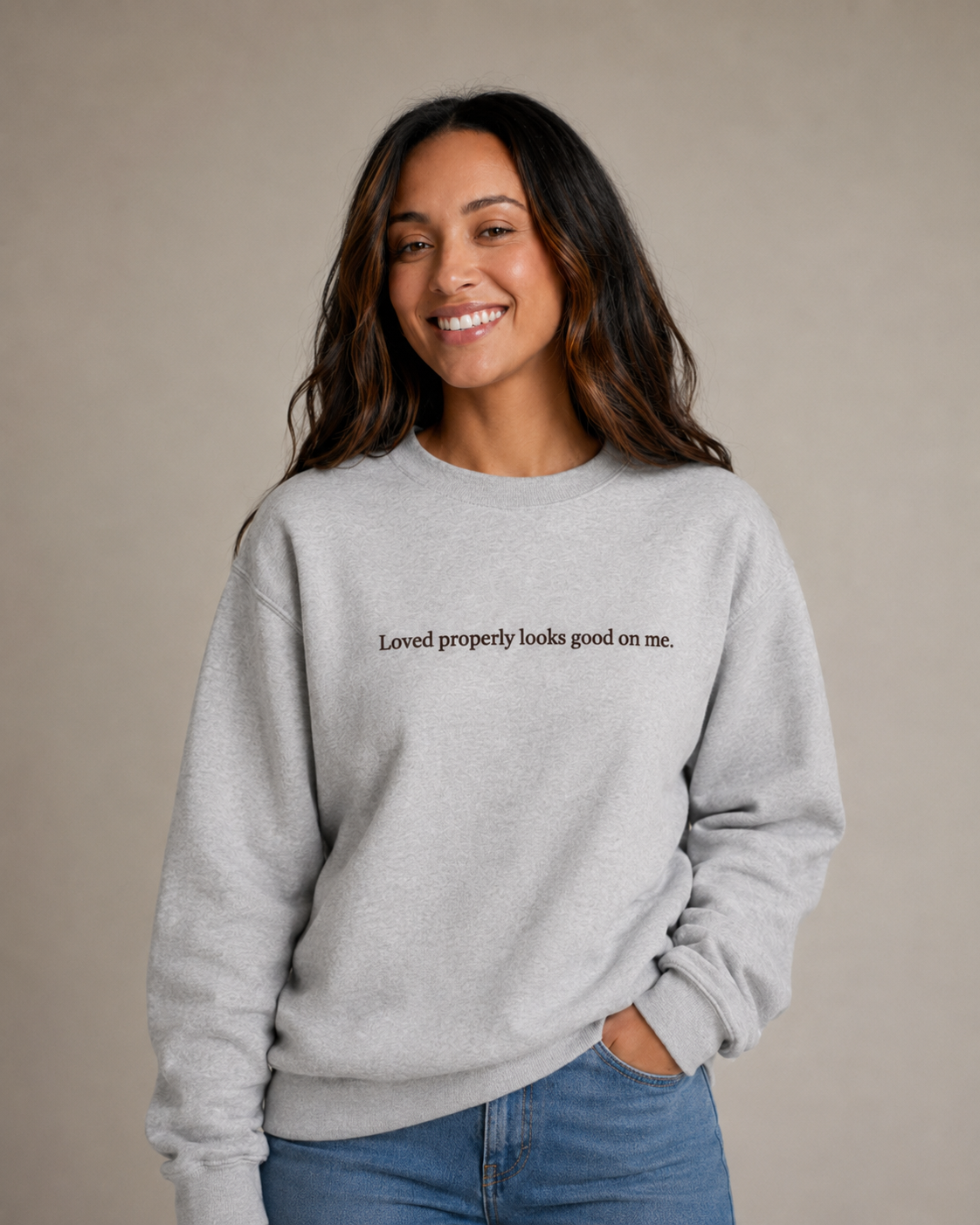 Folded Heather Gray crewneck sweatshirt with dark chocolate embroidery reading “Loved properly looks good on me.” on a light beige fabric background.