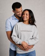 Folded Heather Gray crewneck sweatshirt with dark chocolate embroidery reading “Loved properly looks good on me.” on a light beige fabric background.