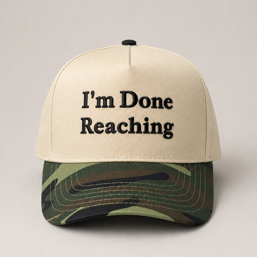A beige trucker-style cap with coffee brown embroidered text reading 'I'm Done Reaching' on the front, and a green camouflage-patterned visor, shown against a plain light gray background.