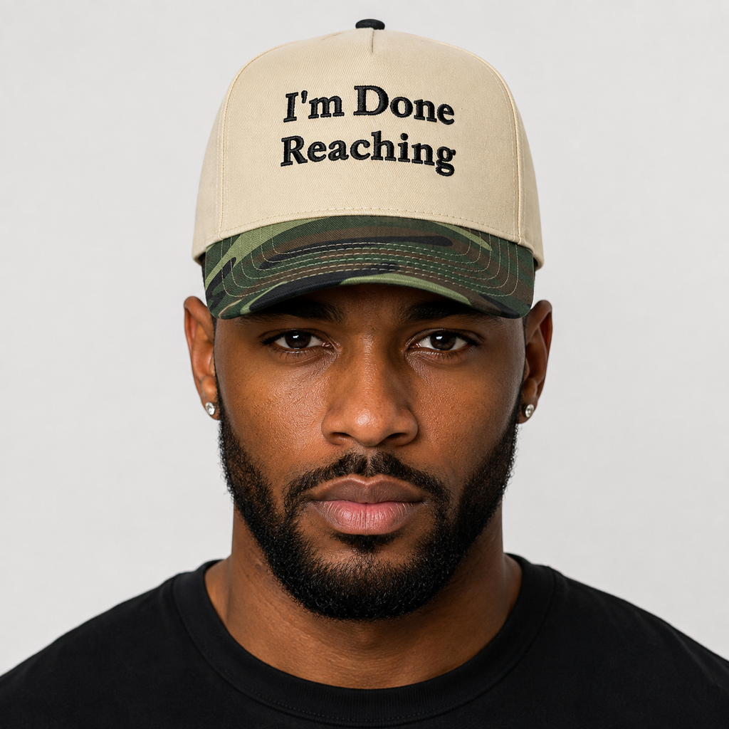 A man wearing a beige baseball cap with coffee brown  embroidered text reading 'I'm Done Reaching' and a green camouflage brim, photographed against a plain light gray background.