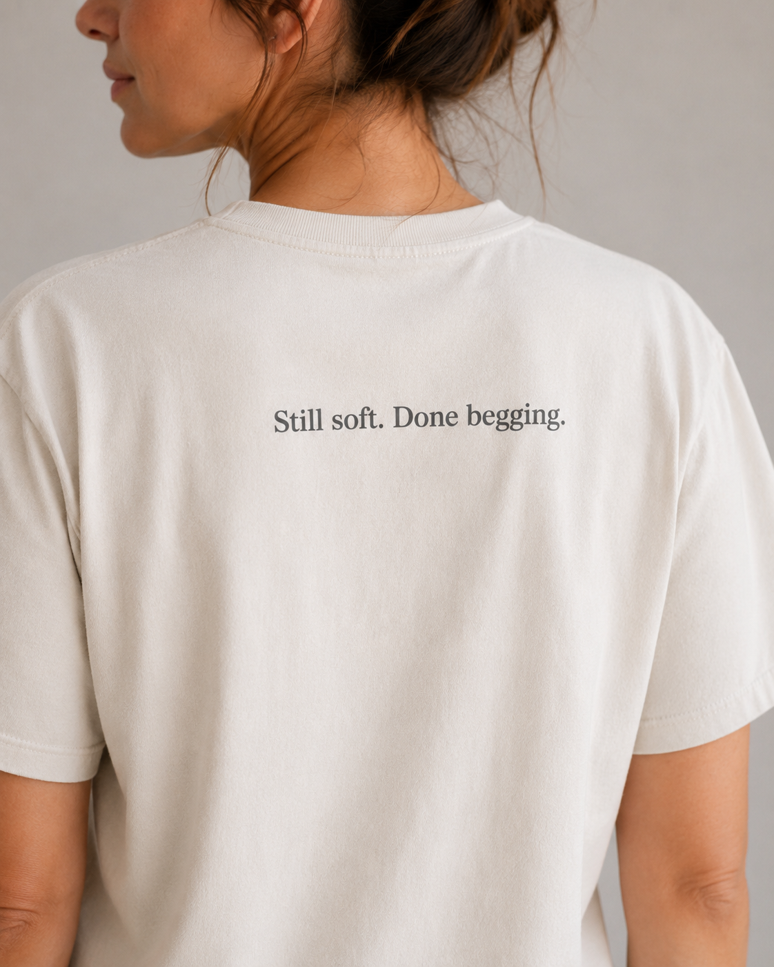 A woman shown from the back wearing a cream-colored t-shirt with the printed text 'Still soft. Done begging.