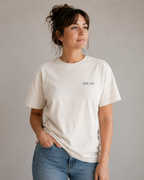 A woman wearing a cream-colored short-sleeve t-shirt with the text 'Still soft.' printed in black on the chest, paired with blue jeans, set against a plain gray studio background.