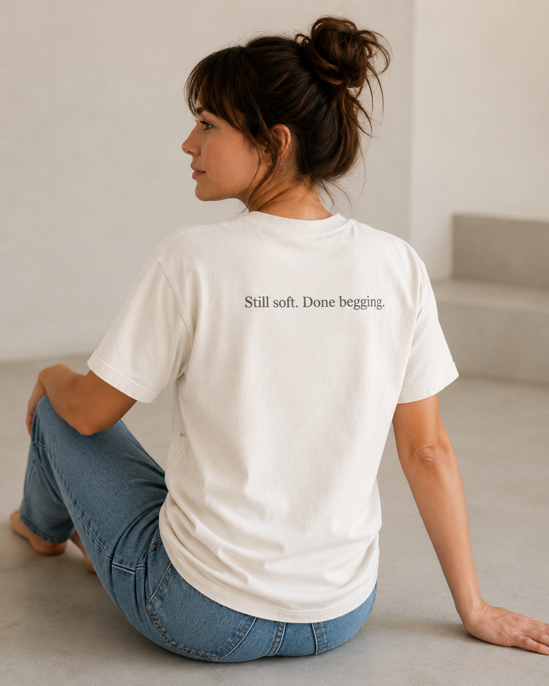 A woman sitting on a concrete floor in a minimalist studio setting, viewed from behind. She is wearing a white t-shirt with the text 'Still soft.