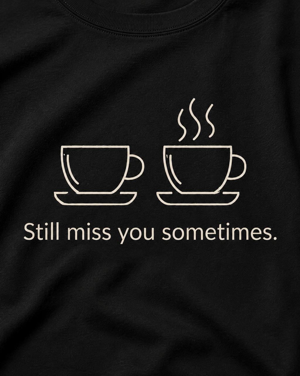 A close-up of a black t-shirt featuring a white graphic of two coffee cups, one plain and one with steam, above the text 'Still miss you sometimes.'.