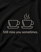 A close-up of a black t-shirt featuring a white graphic of two coffee cups, one plain and one with steam, above the text 'Still miss you sometimes.'.